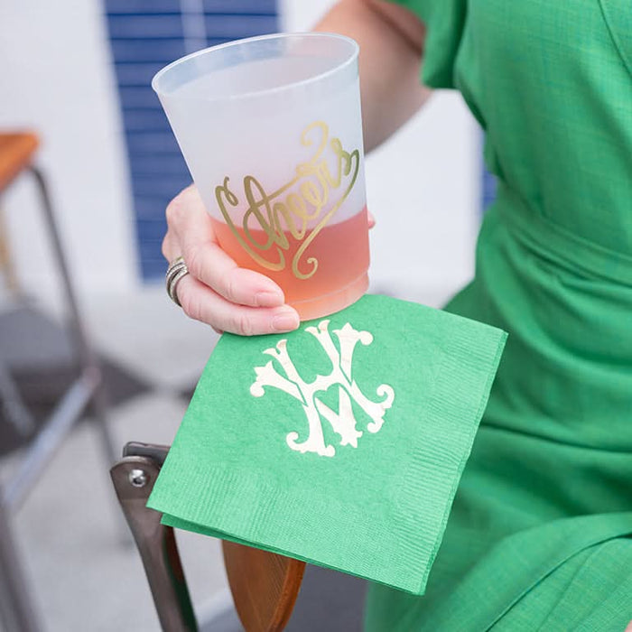 A person in a green dress holds a Cheers 16oz Frosted Cup, with Cheers in gold foil ink, atop a green napkin with a white monogram. They sit by an elegant blue and white tiled wall.