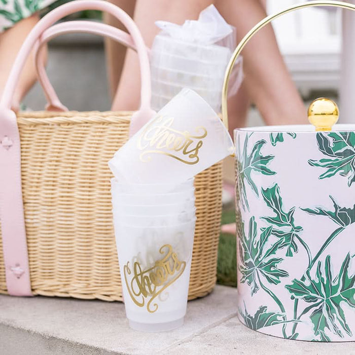 A stack of Cheers 16oz Frosted Cups in gold foil ink rests on a step with a leaf-patterned ice bucket with a gold handle and a straw bag with pink straps. A persons legs are partially visible in the background.