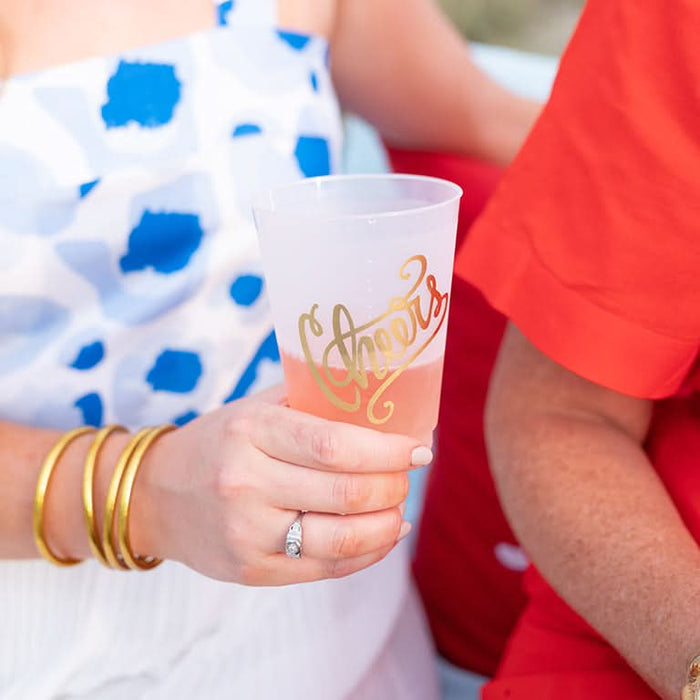 A person in a blue and white patterned dress with gold bangles holds a frosted shatterproof Cheers 16oz Frosted Cup featuring gold foil artwork. The cup contains a pink drink. They sit beside someone in a bright red shirt.