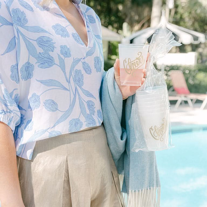 By the pool, someone in a light blue floral blouse and beige pants holds a semi-transparent gift bag containing the Cheers 16oz Frosted Cup Set. A blue blanket is draped over their arm while outdoor chairs and a lush garden complete the scene.