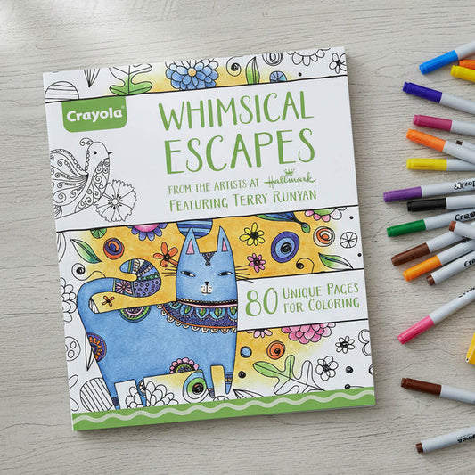 A Crayola 'Whimsical Escapes' coloring book is displayed on a light wood surface next to several colored markers. The book cover features a blue cat with intricate patterns and colorful floral details.