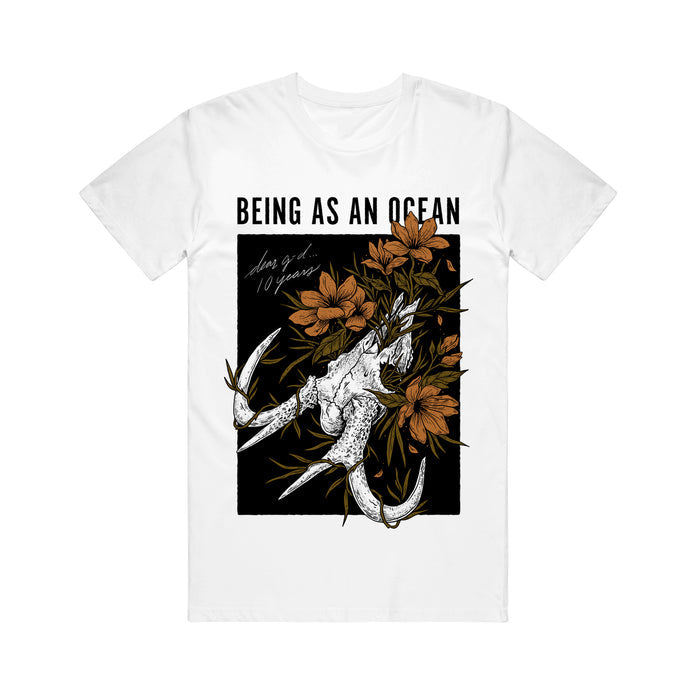 image of a white tee shirt on a white background. the tee has a full body print that says on the top in black being as an ocean with the image of a deer skull below with orange flowers surrounding it.