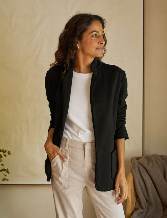 A woman is standing, wearing a black blazer, paired with a white top and beige pants.