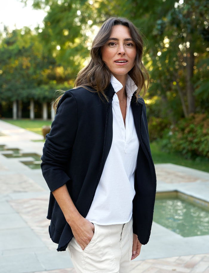 A woman wearing a navy blazer with a white shirt and beige pants standing near a water feature and greenery.