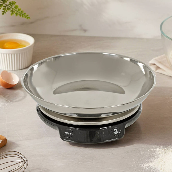 Stainless steel kitchen scales on a worktop with eggs, flour and a whisk