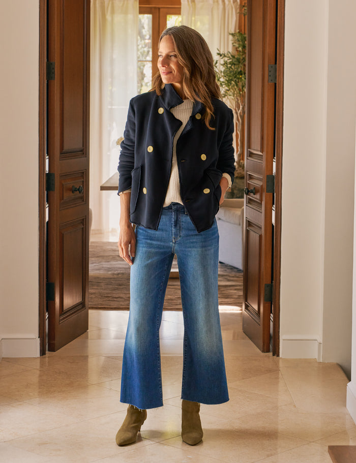 Woman wearing a navy jacket with gold buttons, white sweater, blue jeans, and green shoes in a home setting.