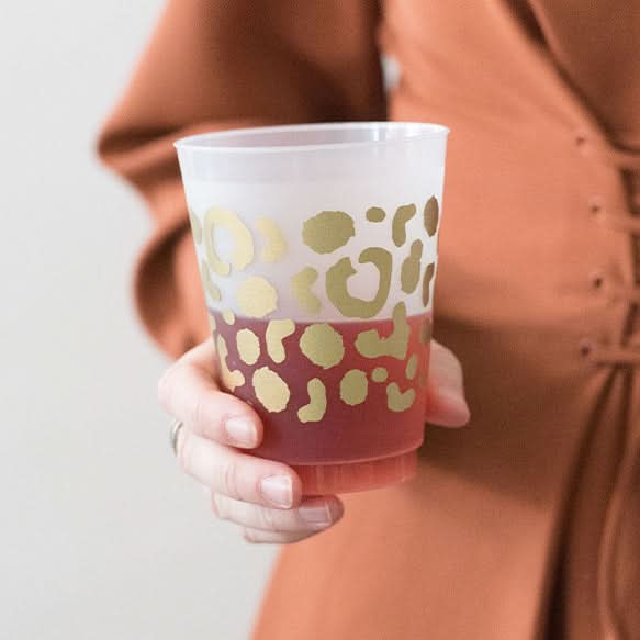 A person holding a Gold Spot Cheetah Frosted Cup filled with a red beverage. The frosted cup boasts a gold leopard print design, akin to a spot cheetah pattern. The individual is dressed in an orange-brown outfit.