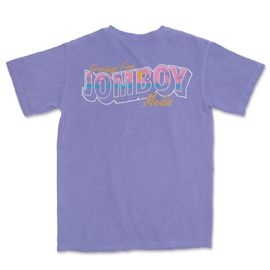 Purple Comfort Colors T-shirt with colorful "Greetings from Jomboy Media" graphic and yellow cursive text.