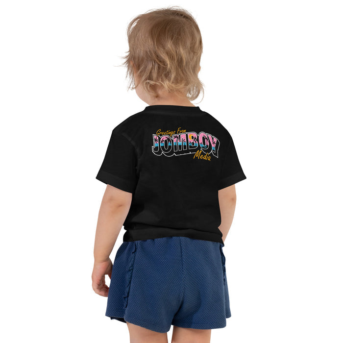 Back view of a toddler wearing a black T-shirt with "Greetings from Jomboy Media" logo and blue shorts.