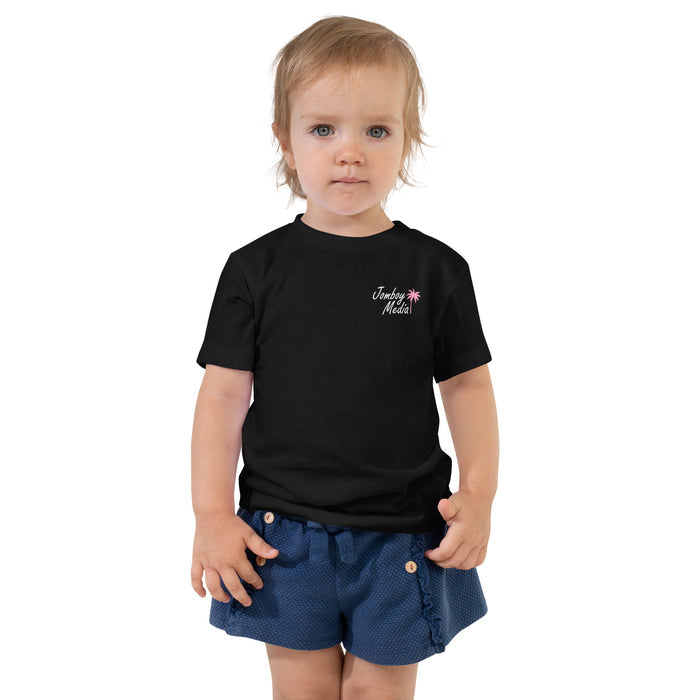 A young child with blonde hair and blue eyes wearing a black "Jomboy Media" toddler tee with pink palm tree logo, paired with blue shorts.