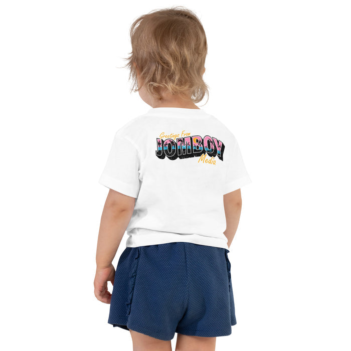Back view of a toddler wearing a white T-shirt with colorful "Greetings from Jomboy Media" text and navy blue shorts.