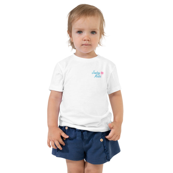Cute toddler girl wearing a white "Greetings from Jomboy Media" t-shirt with a pink palm tree, paired with navy blue shorts.