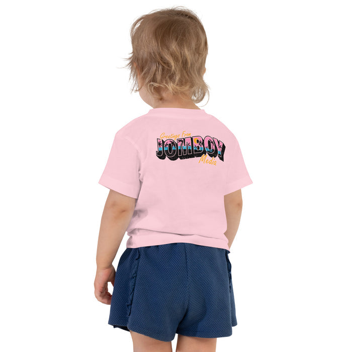 Back view of a young child wearing a pink toddler T-shirt with "Greetings from Jomboy Media" printed on the back, paired with blue shorts.