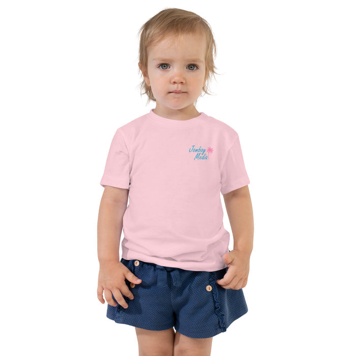 A young toddler girl with blonde hair and blue eyes wearing a pink "Jomboy Media" t-shirt and navy blue shorts, standing against a white background.
