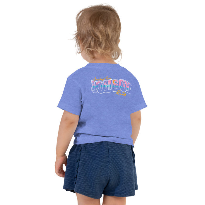 Back view of a toddler wearing a blue T-shirt with "Greetings from Jomboy Media" printed on the back and navy shorts.