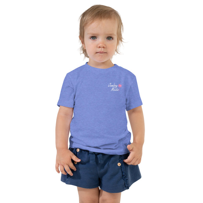 A young child with blonde hair and blue eyes wearing a blue "Jomboy Media" toddler t-shirt and navy shorts, standing against a white background.