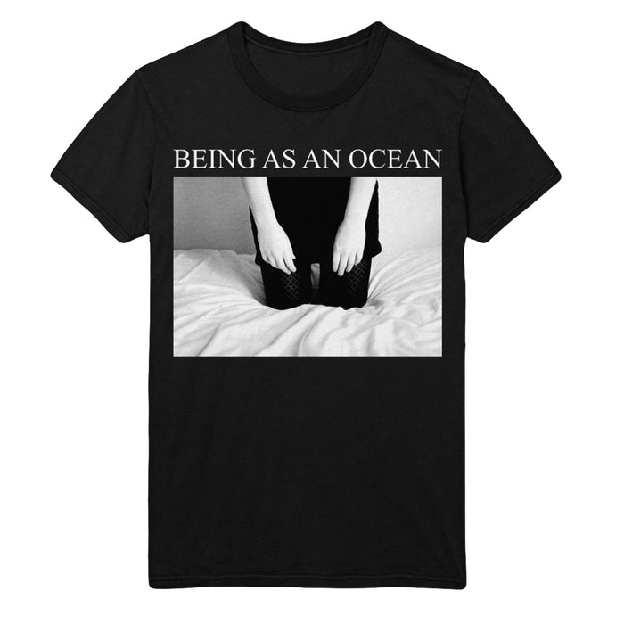 image of a black tee shirt on a white background. tee has a full chest print that says in white on top being as an ocean with a rectangle black and white image of a person kneeling on a bed. the photo is cut off from the forearms down.