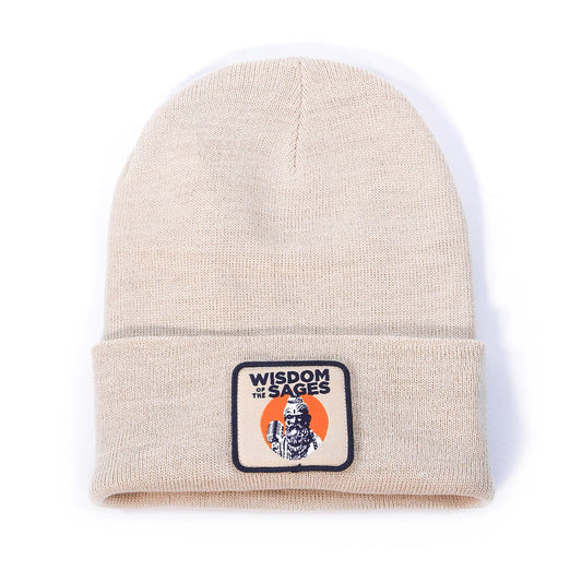 A light beige knit beanie by Wisdom of the Sages, featuring a folded brim with a rectangular patch reading “WISDOM OF THE SAGES” above an illustrated figure and orange semicircle, made from sustainable Polylana polyester.