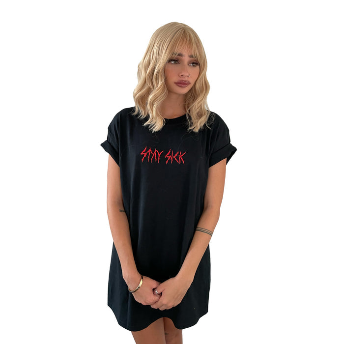 A woman with wavy blonde hair wears a loose black T-shirt dress by Stay Sick Clothing, featuring red metal-embroidered SAD SICK lettering, standing against a white background with her arms at her sides.