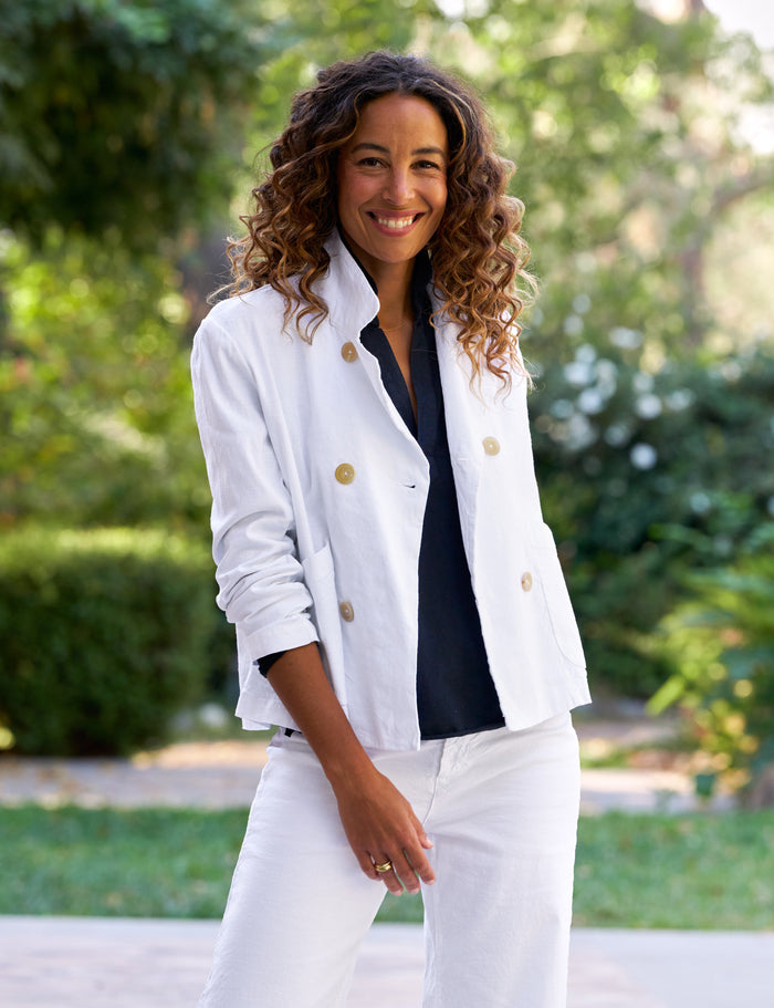 Woman wearing a white jacket with brown buttons and a navy shirt, standing outdoors with greenery in the background.