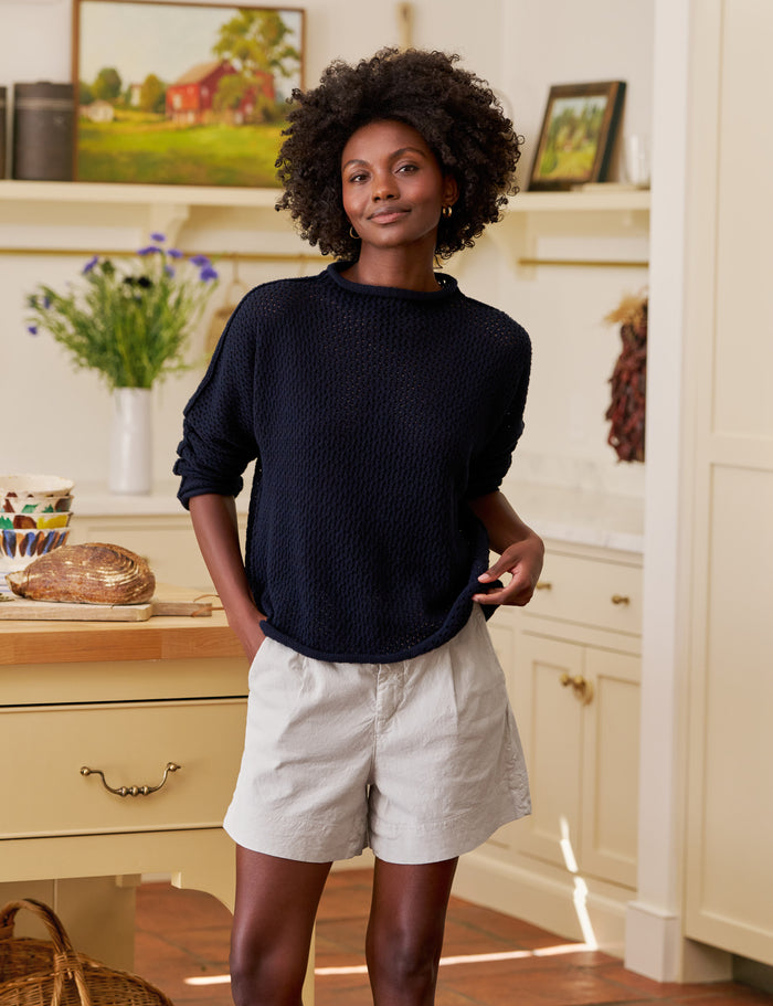Woman standing in a kitchen wearing a navy sweater and light shorts.