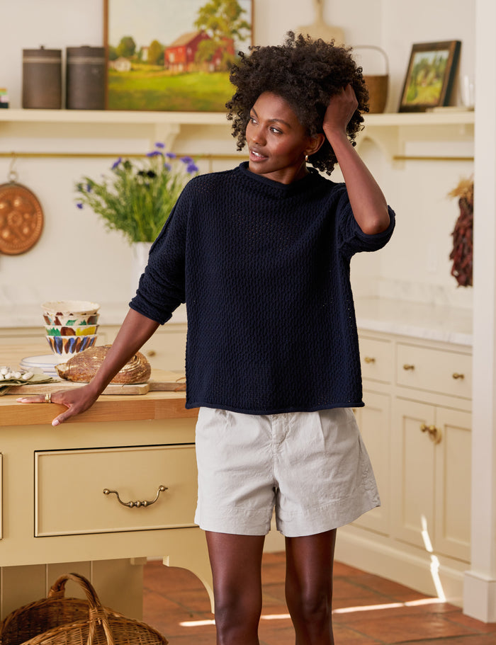 Woman standing in a kitchen wearing a navy sweater and tan shorts.