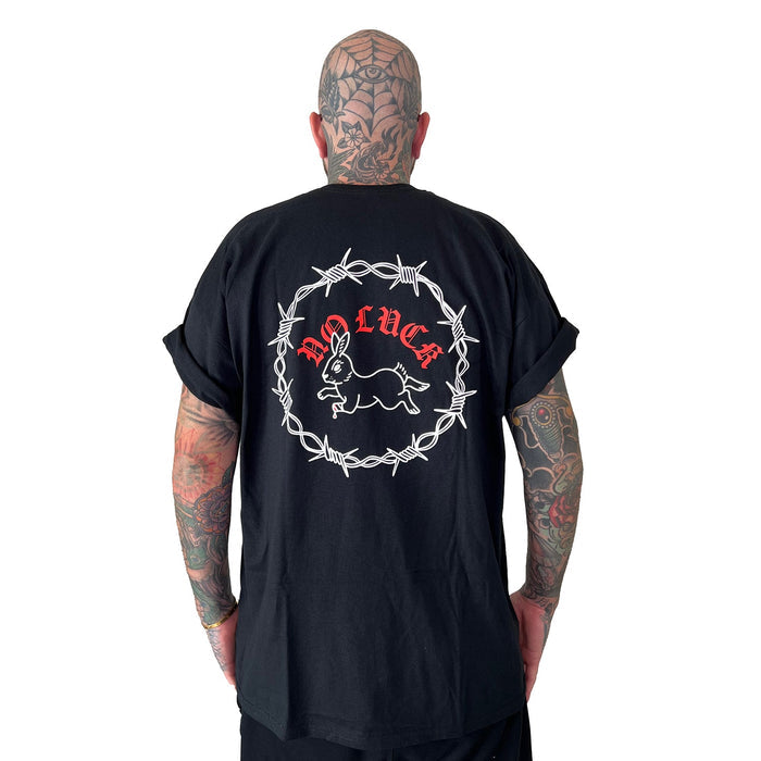A tattooed individual faces away, wearing a Stay Sick Clothing Black T-Shirt. The shirt’s back features a white barbed wire circle, rabbit illustration, and bold red NO LUCK text, all set against a plain white background that highlights the striking design.