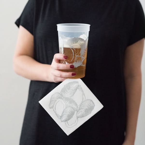 A person in a black shirt holds a translucent cup of iced tea with an octopus design, paired with Oyster Cocktail Napkins in white and silver.