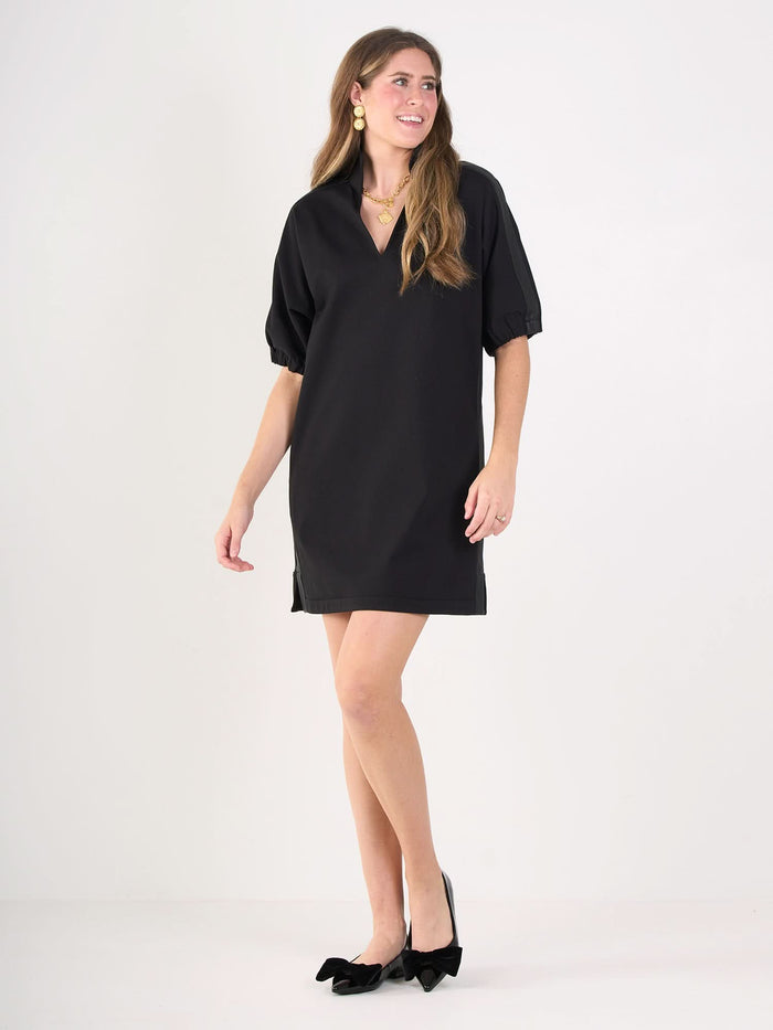 A woman smiles while standing against a plain white background, wearing the Poppy Dress—a loose-fitting short black ponte dress with elbow-length sleeves—paired with black bow heels and gold jewelry.
