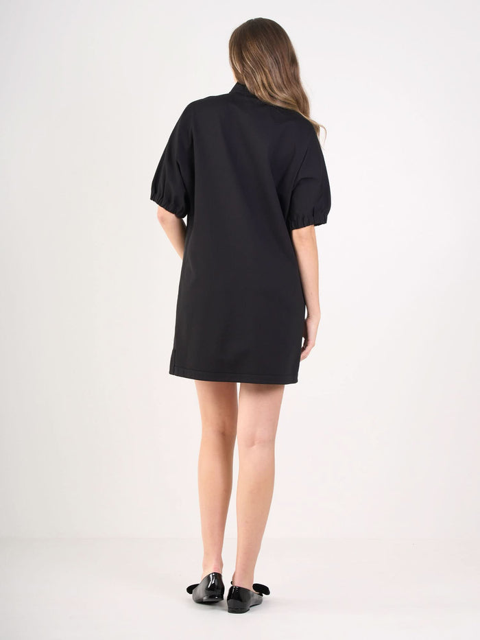 A woman with long, wavy hair faces away, wearing the Poppy Dress—a short black dress with puffed sleeves—and black flats, standing against a plain white background.