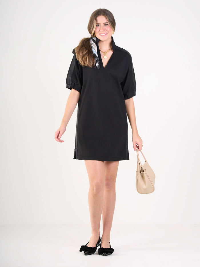 A woman stands smiling in a Poppy Dress with short sleeves and a deep collar, wearing black shoes with bows, a patterned hair scarf, and holding a beige vegan leather handbag against a plain white background.