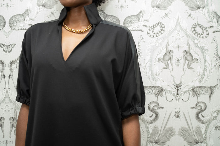 A person in a Poppy Dress - Black Ponte, adorned with a gold chain necklace, stands before an intricately patterned wallpaper featuring animals and foliage. The V-neckline and half-length sleeves emphasize their style, while their face remains unseen in the image.