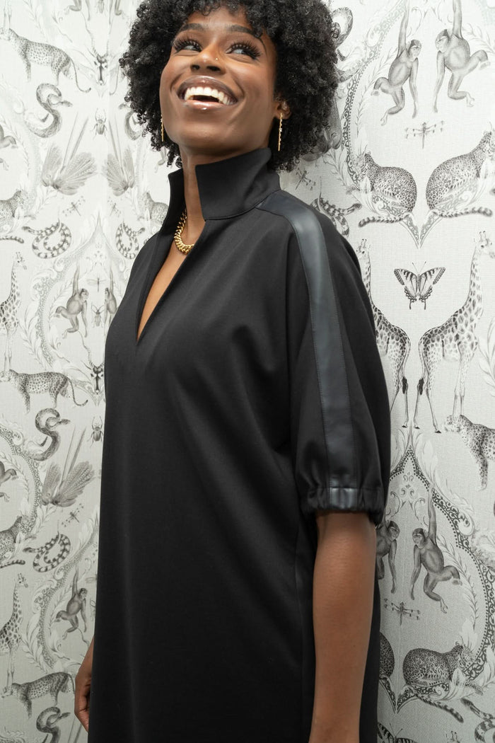 A smiling woman in the Poppy Dress, featuring black fabric and vegan leather shoulder stripes, stands before wallpaper decorated with illustrated animals and plants.