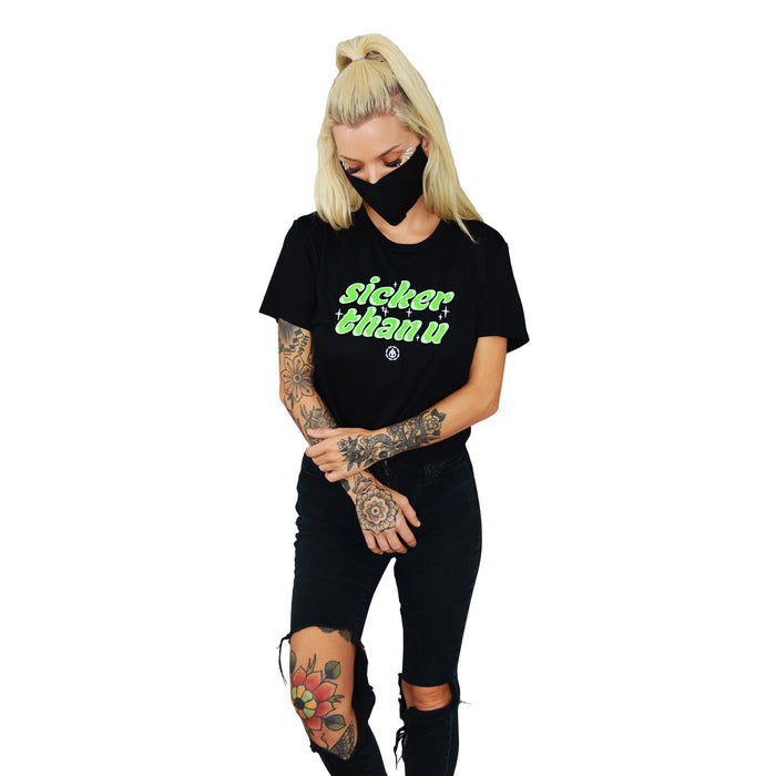 A woman with blonde hair in a high ponytail wears a Stay Sick Clothing black cropped T-shirt with “sicker than u” in neon green, ripped black jeans, and a mask. Tattoos on her arms and leg add to the edgy vibe—perfect for your Black Girl’s T-Shirt collection.