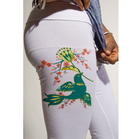 A person wearing Mantralogy white yoga pants with a fold-over waistband and a colorful green hummingbird and flower design on the upper thigh, with their torso and hand partially visible.