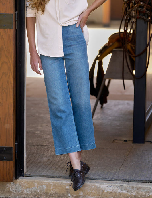 A person wearing wide-leg cropped blue denim jeans, a cream blouse, and dark brown leather shoes, standing in a doorway.